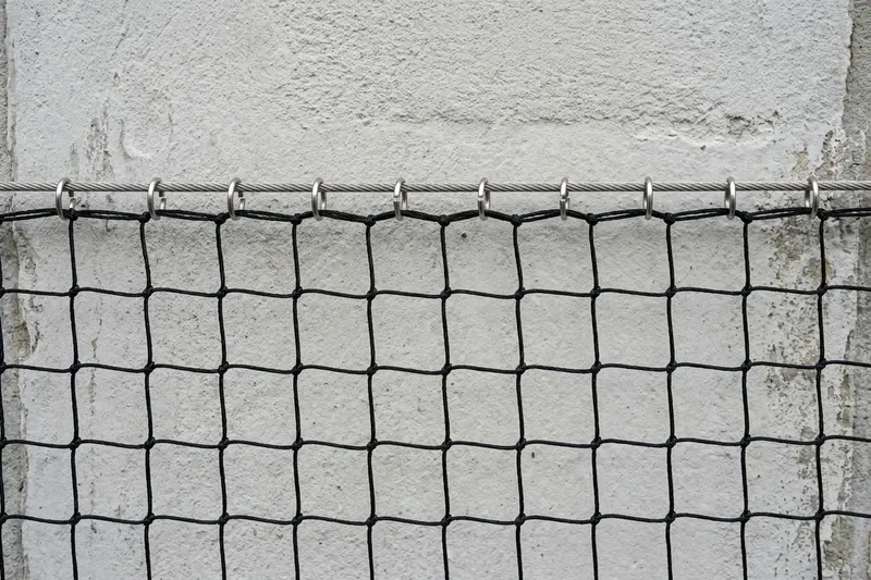 Närbild av en fågelvajer längs en putsad fasad där nätets kantmaskor är fästa med små metallringar, en ring per maska
