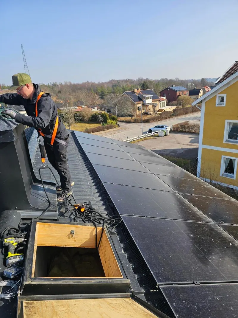 Tekniker i fallskyddssele och keps arbetar på ett tak med solpaneler, med villor och träd nedanför